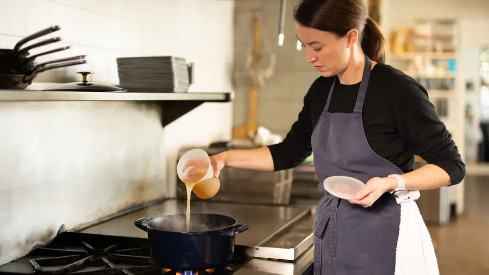 A focused chef pours liquid ingredients into a pot on a commercial kitchen stove, indicating a cooking process in action.