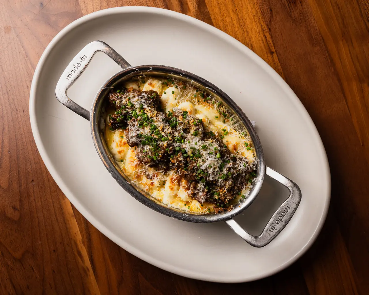 A baked dish featuring tender meat topped with a crispy breadcrumb layer and herbs, served in a metal dish over creamy cheese.