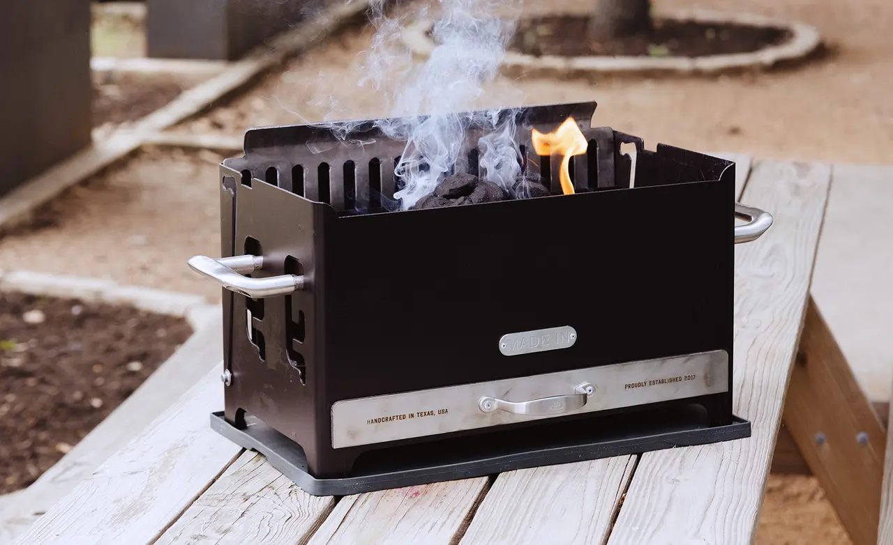 A portable charcoal grill sits on a wooden surface, emitting smoke and a small flame.
