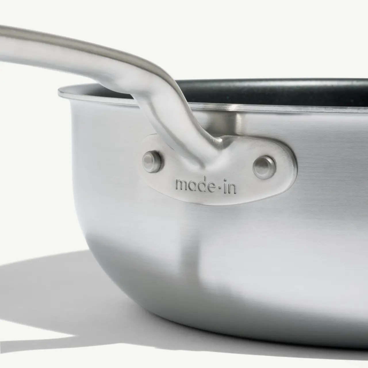 A close-up of a stainless steel saucepan with the text "made in" etched near the handle, highlighted by soft lighting casting a gentle shadow.