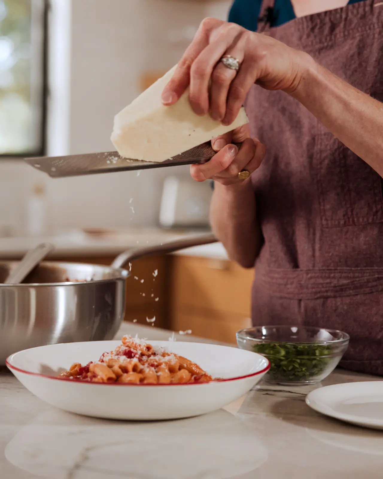 A person in an apron is using a knife to grate cheese over a bowl of pasta, with a pot and herbs visible in the background.
