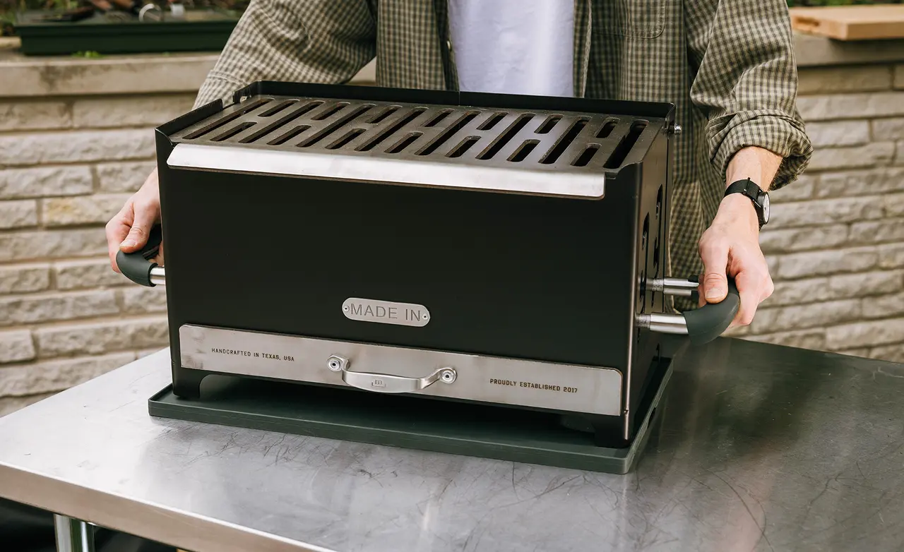 A person is holding a black and stainless steel stovetop grill with the label "Made In," positioned on a stainless steel table.