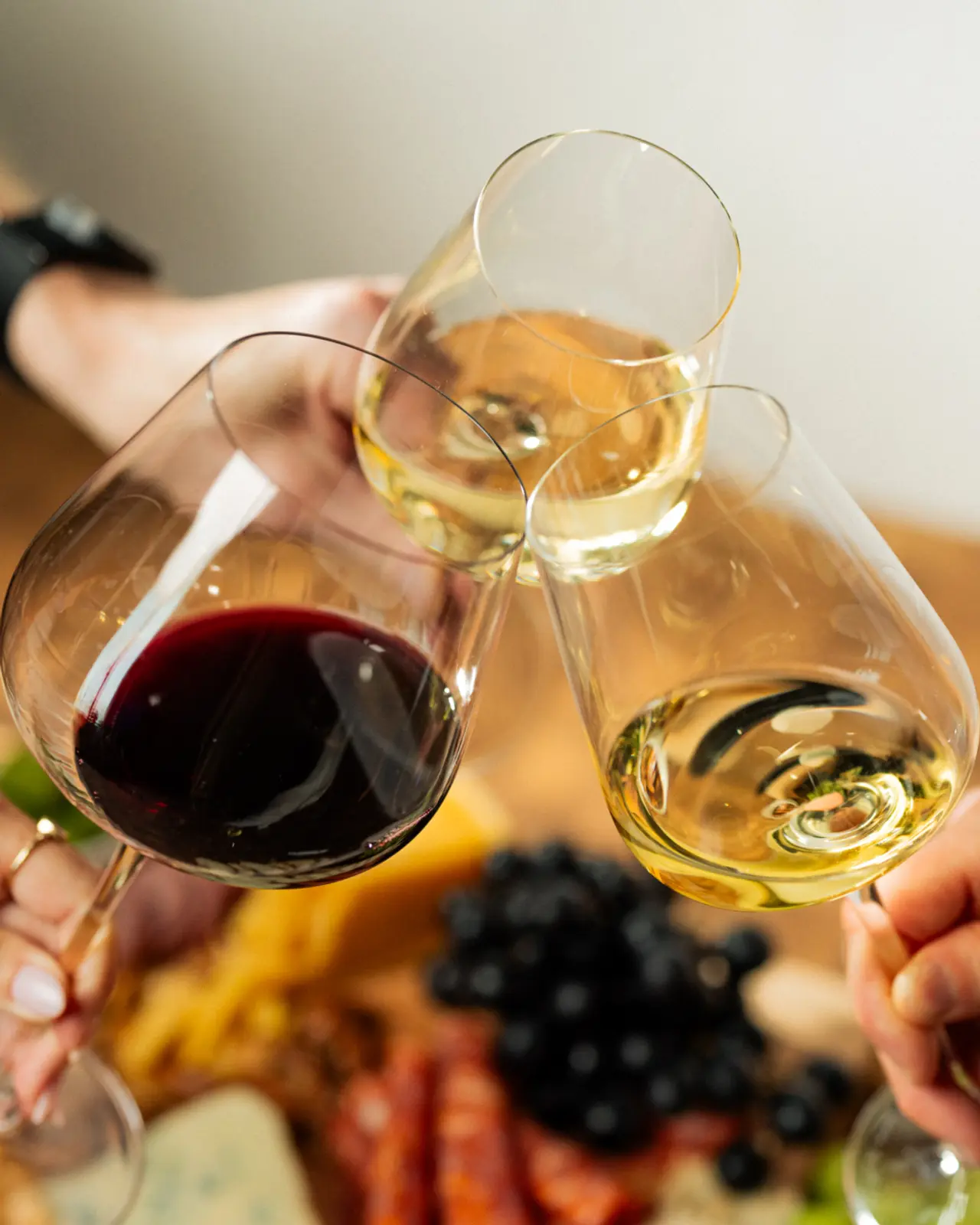 Hands are toasting with glasses of red and white wine above a table laden with grapes and charcuterie.