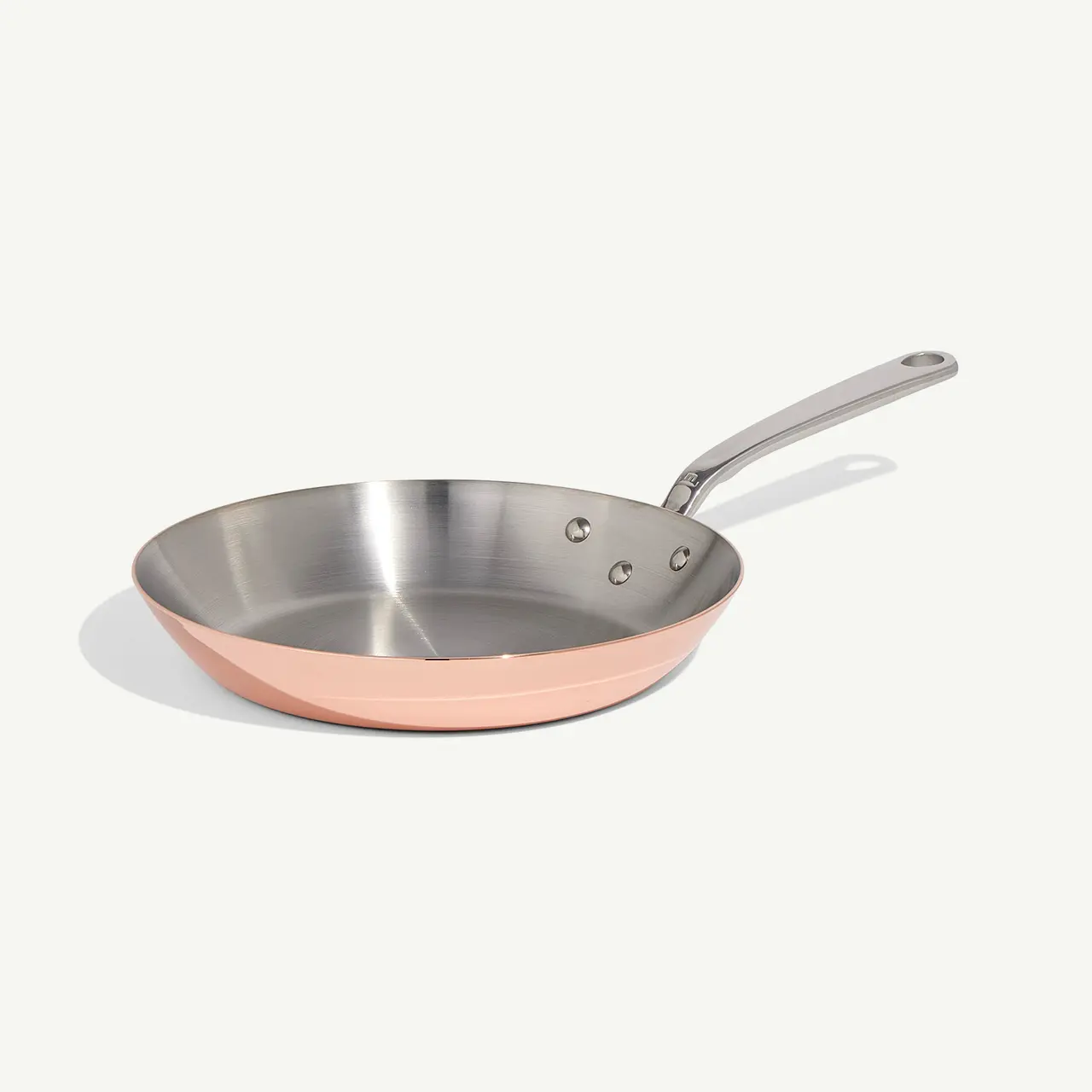 A stainless steel frying pan with a copper exterior and a long handle rests on a light background.