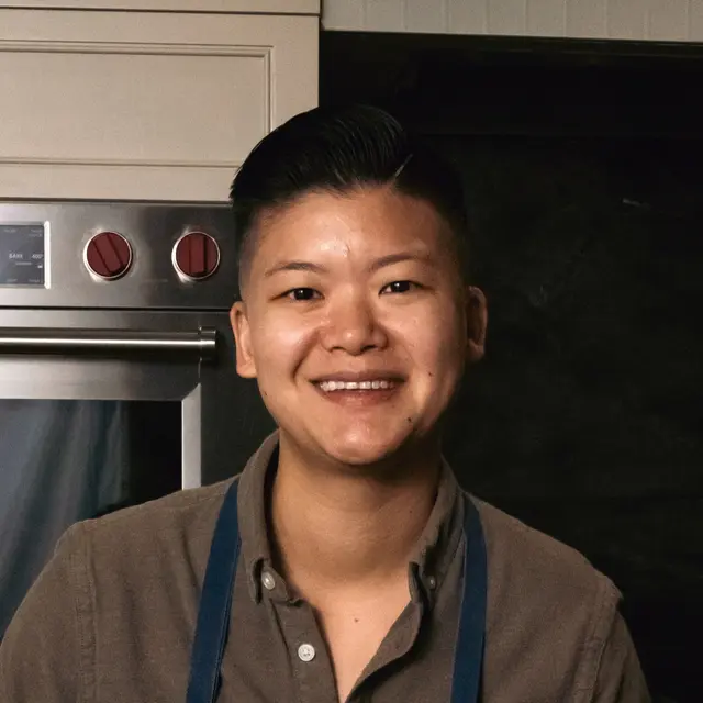 A smiling person with a styled haircut wearing a gray shirt and a blue apron stands in a kitchen setting.