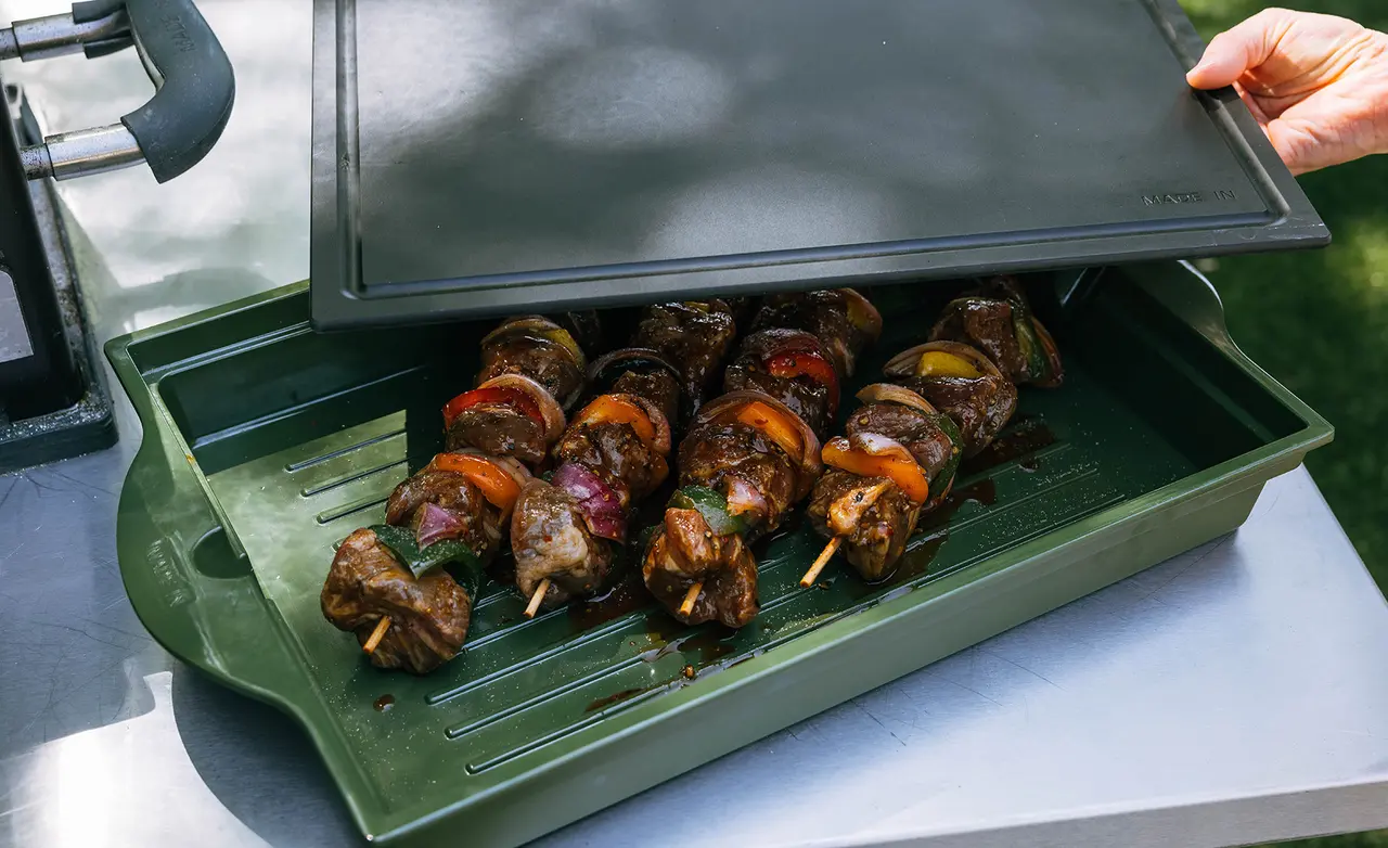 Grilled kebabs with vegetables are being lifted from a rectangular grill container with a lid.