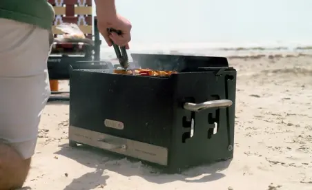 A person is grilling food on a portable barbecue near the beach, with the ocean visible in the background.