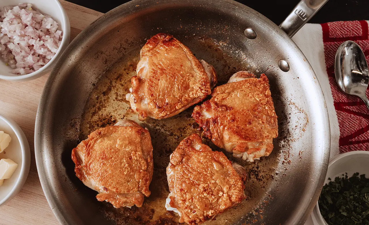 Four golden-browned chicken thighs are sizzling in a stainless steel skillet next to small bowls of shallots, butter, and chopped herbs.