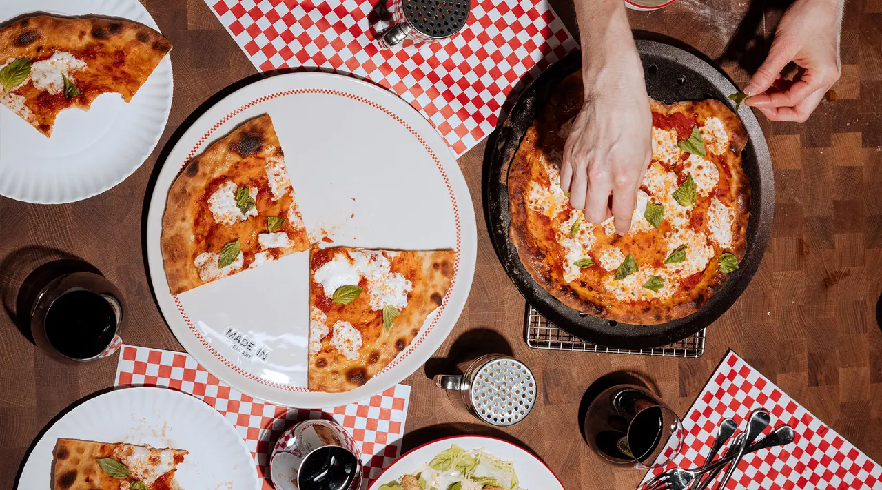A person is reaching out to take a slice of pizza from a pan placed on a table set with plates, glasses, and utensils, with another pizza already partially sliced.
