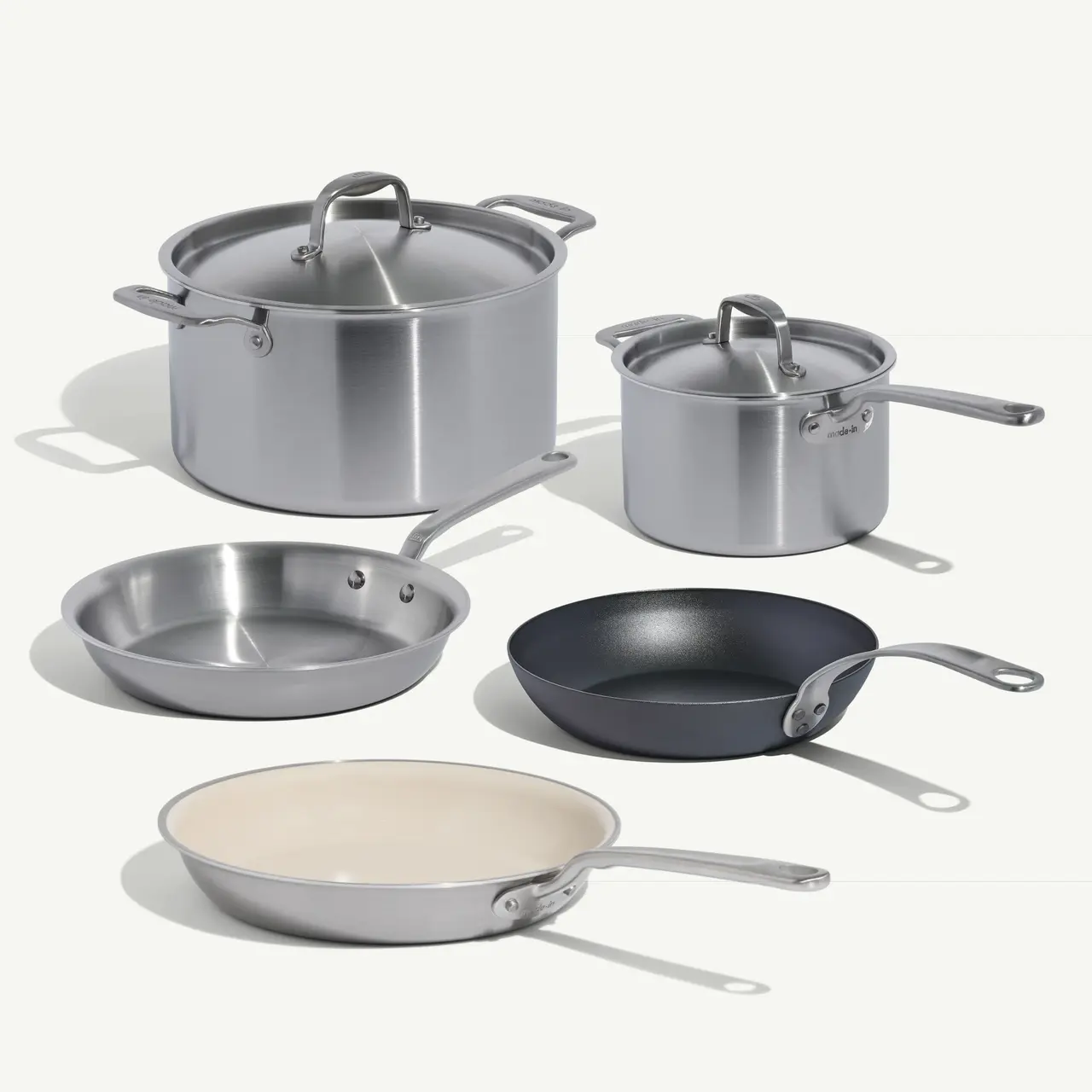 A collection of five cookware pieces, including stainless steel pots and pans in various sizes, is arranged on a light background.